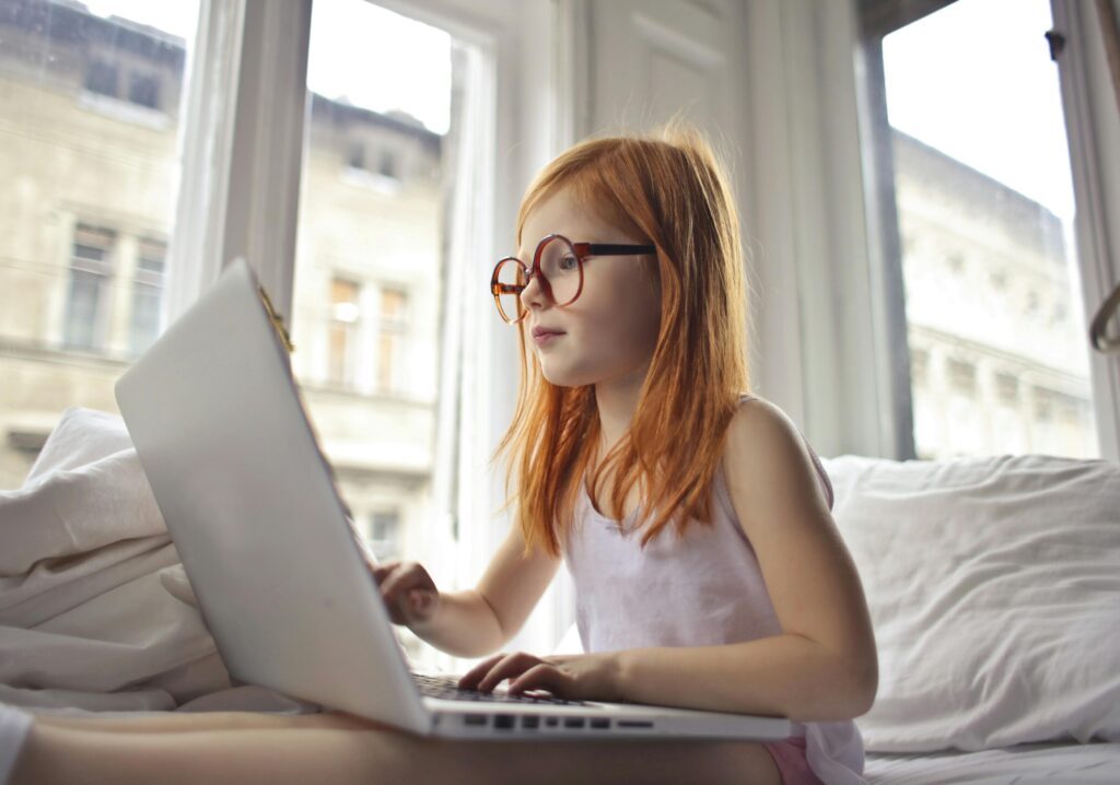 Young girl using a laptop in a sunny bedroom. Perfect for themes of tech and leisure.